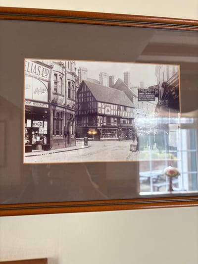 A framed, monochromatic photograph depicts a bustling street scene in West Felton, United Kingdom, featuring historic architecture and pedestrian activity. The central focus is a prominent half-timbered building with a gabled roof and multiple windows, its dark wooden beams contrasting with lighter infill, located at a street intersection.

To the left of this central building, a multi-story structure with ornate stonework and bay windows displays prominent signs for "MELIAS LTD" and "PROVISION MERCHANTS." Its ground-level storefront exhibits various goods. A street object, possibly a post box or bollard, stands on the pavement in front of the half-timbered building.

To the right, another building features multiple commercial signs, including "THE CROSS," "FOSTER BROS," "EDWARD THOMAS & C," "GENERAL IRONMONGERS," "IMPLEMENT MERCHANTS," and "MILITARY STORES." Several indistinct figures, appearing as pedestrians, are visible on the pavement further down the street on the right. The street itself appears paved, with a visible curb line.

The photograph is contained within a brown wooden frame with a dark grey mat. Reflections on the frame's glass obscure portions of the image, most notably a window frame and a patterned table lamp on the right side, and a blurred, dark reflection on the left. Faint text in the bottom left of the print reads "--- to Hapleston" or similar, while the number "49504" is visible in the bottom right.