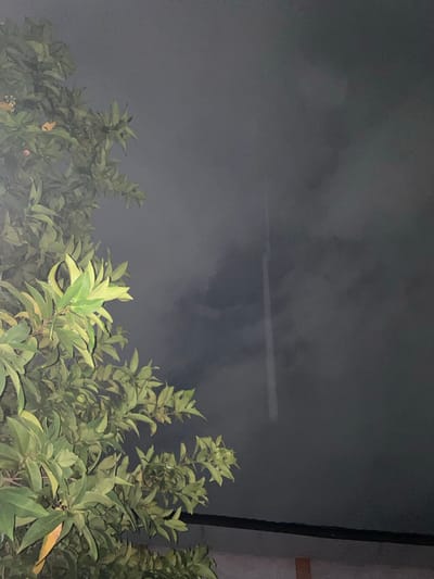 A nighttime photograph captures a close-up view of lush green foliage from a tree in the foreground, which occupies the left side of the frame. The leaves are well-lit, appearing vibrant and detailed against the dark, hazy sky. In the upper right quadrant of the image, a faint, vertical white line is visible, possibly a light streak or a distant object. The bottom edge of the image reveals a dark, horizontal band, likely a roof or structure, with a sliver of a lighter-colored wall beneath it. The overall atmosphere is dark and somewhat mysterious, with the bright foliage contrasting against the muted sky. The scene is identified as being located in Banjarmasin, Indonesia.