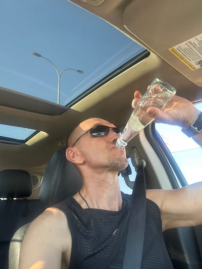 A bald man wearing sunglasses and a black tank top is drinking from a glass bottle of water while driving a car. The sun is shining through the sunroof, illuminating the interior of the car. The man is in the driver's seat, and the passenger seat is visible behind him. The car appears to be parked on a street in Omer, Israel, as evidenced by the streetlights visible through the sunroof.