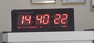 An interior static image depicts a black rectangular "Numeral message Calendar" digital display unit, showing red LED segment text. The unit displays the time as 14:40:22, the date as 18 Day 1 Mon 2026, and the temperature as 25 °C. The day of the week "SUN" is illuminated. The digital unit rests on a flat, white surface, likely a shelf or desktop. In the background, a plain, light-colored wall is visible, on which two framed items are mounted. The left frame is gold-colored, enclosing a light-colored document. The right frame is darker, also containing a document or image. The scene is located in Kogon City, Uzbekistan.
