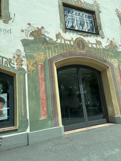 The image captures the facade of a building in Reutte, Austria, characterized by elaborate fresco paintings. The artwork depicts classical architectural elements such as columns and decorative borders, painted in shades of green, gold, and red. There are floral motifs with red roses and green foliage. A prominent arched doorway with double glass doors is visible, reflecting the surrounding trees. Above the doorway, a window with ornate black metal bars is set into the wall. To the left of the doorway, a window displays a poster of a person's head, suggesting a business such as a hairdresser. The building's exterior is a light color, possibly stucco, with the painted decorations significantly covering its surface. The ground in front of the building is asphalt. The overall impression is of a historic building adorned with traditional artistic embellishments.