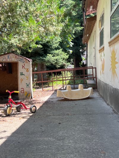 The image depicts an outdoor area, possibly a playground or yard, in Akandzhievo, Bulgaria. A small red tricycle with yellow handlebars is positioned on the left side of the frame, casting a shadow. To the right of the tricycle, a beige plastic seesaw is situated on the asphalt ground. Behind the tricycle, a small, shed-like structure is visible, decorated with colorful letters and a butterfly. A metal fence and lush green trees form the background. The building on the right side of the image has a white facade adorned with painted suns and other decorative elements. The overall scene appears to be a sunny day.