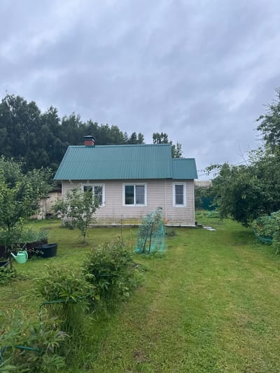 The image depicts a residential house with a green metal roof and beige siding, situated in a rural environment. The house has two visible windows on its front facade and a chimney with a red cap. The surrounding area is a grassy yard with various plants, bushes, and trees. There are also gardening accessories such as a green watering can and a black tub. The sky is overcast. The location is identified as Красное, Russia.