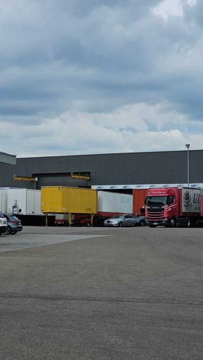 The image depicts an outdoor scene in Neu-Ulm, Germany, likely a logistics or industrial area, under a cloudy sky. In the foreground, a gray asphalt surface forms the ground, with tire tracks visible. Parked to the left are two silver cars. Behind these cars are several shipping containers and trailers. A bright yellow trailer with two axles is positioned centrally. To its right, a white container with red accents and the text "WÖRGEL KRAFT" is visible. Further to the right, a red Scania truck with the branding "Ulrich Hinz" is parked, connected to a white and red trailer. The truck cab displays a license plate starting with "UL". The background features a long, gray, corrugated metal building with multiple entrances or loading bays, one of which appears to be open with a yellow gantry crane partially visible. Several white trailers with logos, including "VTI" repeated multiple times, are lined up in front of the building. The overall atmosphere is industrial and functional.