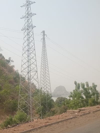 The scene is set in Jebba, Nigeria, under a hazy, light gray sky. Two galvanized steel lattice transmission towers dominate the midground. The primary tower, taller and wider, is positioned to the left, while a second, thinner tower stands further back and to the right. Multiple power lines extend horizontally across the upper portion of the frame from these structures.

To the left of the foreground tower, a steep hillside covered in dry brush and scattered green trees ascends. A body of water, consistent with the River Niger, is visible between and behind the towers, its distant bank lined with dense green foliage. In the far background, a distinctive, rounded rock formation, characteristic of Jebba, rises from the water, partially obscured by atmospheric haze.

The immediate foreground features a dirt road shoulder, light brown in color, with a concrete curb delineating the edge of a paved road. Scattered concrete debris is present on the shoulder. No individuals or discernible actions are depicted.