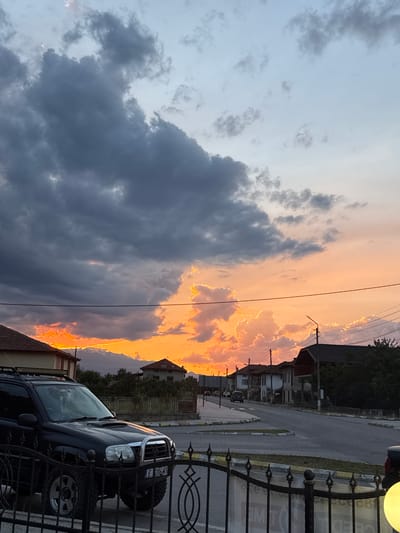 This media file captures a dramatic sunset over a residential area in Blatska, Bulgaria. The sky is a canvas of vibrant colors, with fiery oranges and yellows illuminating the edges of dark, heavy clouds. The light from the setting sun casts a warm glow across the scene, highlighting the textures of the clouds.
In the foreground, a black SUV is parked behind a decorative metal fence. The vehicle has large, rugged tires and a chrome bumper, suggesting it might be an off-road vehicle. The fence has intricate scrollwork and posts, and a portion of a yellow outdoor light fixture is visible at the top right.
Behind the fence and the SUV, a street runs through the neighborhood, lined with houses and trees. The architecture of the houses appears traditional, with pitched roofs. Power lines are visible stretching across the sky, and a lamppost stands along the side of the road.
The time of day is clearly sunset, given the intense coloration of the sky. The weather appears to be clearing, with the dark clouds possibly indicating recent rain or an approaching storm, contrasted with the bright, sunny sky on the horizon. The overall mood is one of natural beauty and the transition from day to night.
Visible text on the license plate of the SUV reads "E 206 MT". There is also some partially obscured text on a sign behind the fence, possibly advertising or informational.
