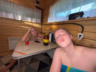 Two young women are posing for a selfie in a wooden room, possibly a sauna or a rustic cabin. The room is lined with wooden planks. The woman in the foreground is closer to the camera, her face close to the lens, with her eyes closed and lips pursed in a kiss. She is wrapped in a blue and yellow towel. The woman in the background is sitting at a small table, holding her head with one hand and looking at the camera with wide eyes. She has a glass of red liquid, a bottle of brown liquid, and a smartphone on the table in front of her. A white electrical outlet is visible on the wooden wall behind the woman in the foreground. A digital clock is mounted on the wall above a window covered by white curtains. The lighting suggests it is daytime, and there are no obvious indications of the weather outside. The overall mood seems playful and relaxed. The label on the brown bottle appears to be in Russian and says "ЖЕЛТАЯ РЕКА" which translates to "Yellow River".