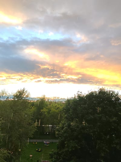 This media file captures a sunset with a cloudy sky, painted in hues of orange, yellow, and blue, with some streaks of white and gray clouds. The sun is setting, casting a warm glow across the sky, suggesting it is late evening. The scene transitions to a lush green landscape filled with trees, creating a natural setting. In the foreground, dense foliage of trees dominates the view, partially obscuring the background.

Beneath the trees, a grassy area is visible, featuring a playground with colorful elements like a red and blue airplane-shaped structure, yellow chairs, and a yellow and black toy. A white picket fence surrounds part of the area, and hints of other play equipment are discernible. The presence of a playground suggests a residential or park area.

The weather appears to be somewhat unsettled, with visible streaks that could indicate falling rain or perhaps a visual artifact in the image. The overall mood is serene and tranquil, amplified by the soft lighting of the sunset. The location is identified as Sokol, Russia. There is no visible text in the media file.
