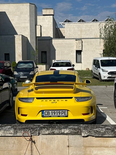 In Arbanassi, Bulgaria, a yellow Porsche GT3 is parked in a lot in front of a modern stone building. The car is facing away from the camera, with its rear visible. The building has a light-colored stone facade and geometric architectural elements, with some windows and shadowed recesses. Several other vehicles are parked around the Porsche, including a white van, a white Audi, and a dark-colored hatchback. The sky is blue with scattered clouds. A stone wall with a chain and rope is in the foreground.