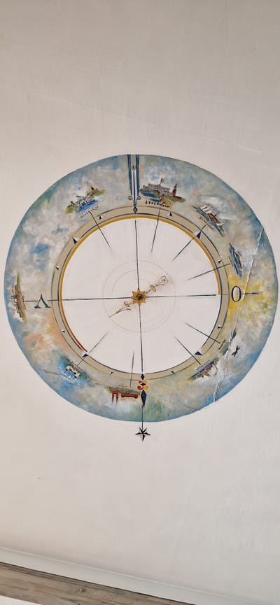 The image is a ceiling mural depicting a compass rose with various Swedish cities and landmarks around its circumference. The mural is painted in a realistic style with a muted color palette, featuring blues, whites, grays, and touches of yellow and brown. The central part of the compass rose has a decorative metal clock hand pointing towards the upper left.

Around the outer edge of the compass, small vignettes of different locations are painted, each accompanied by its name in capital letters. Visible text includes: "MÖJA," "ARLANDA," "NORRTÄLJE," "UPPSALA," "VÄSTERÅS," "SÖDERTÄLJE," "STOCKHOLM," and "SKÄRHAMN." Each vignette depicts characteristic scenery, such as buildings, boats, airplanes, or natural landscapes, suggesting a maritime or travel theme. For example, "STOCKHOLM" shows a cityscape with prominent buildings, and "ARLANDA" features an airplane. The overall impression is a stylized representation of geographical locations connected by a compass, possibly indicating routes or directions. The mural is painted on a white, textured ceiling, and the lighting suggests it's daytime.