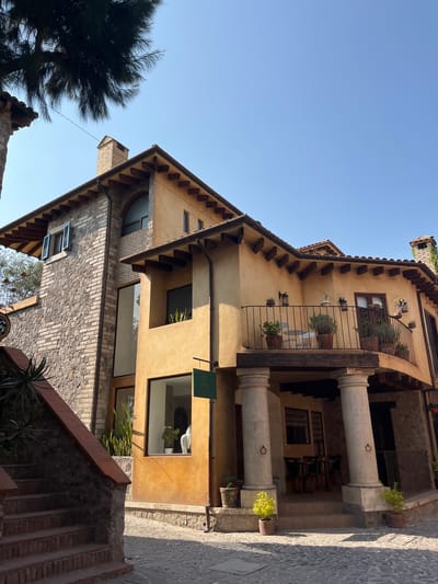 The image captures a sunlit architectural scene in Domingo Arenas, Mexico, featuring multi-story buildings with a blend of stone and stucco facades. The architectural style exhibits elements of Mediterranean or Tuscan design, characterized by exposed beams, tile roofs, and arched balconies with potted plants. Stone steps and pathways delineate the foreground, while the clear blue sky offers a backdrop to the buildings. The scene appears tranquil, devoid of human activity, and emphasizes the architectural details and natural light.