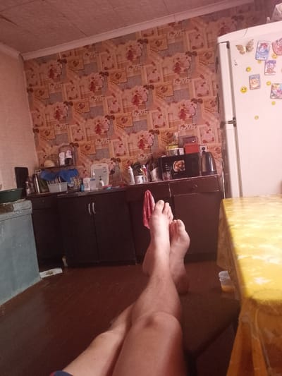 The image depicts an interior scene, likely a kitchen, with a focus on a person's legs and feet positioned in the foreground. The lower body of a person is visible, with their legs crossed. Their feet are bare and appear to be resting. Behind the legs, a kitchen area is visible. This includes a countertop with various items such as bottles, a sink, and a microwave. A refrigerator is also present on the right side of the image, adorned with magnets. The walls are covered with patterned wallpaper. The flooring appears to be made of wood. A table with a yellow tablecloth is partially visible on the right. The overall lighting suggests an indoor environment, possibly illuminated by artificial light. The scene is devoid of any explicit indicators pointing to the city of Ivanovskaya, Russia.