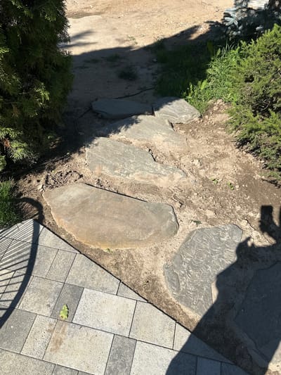 The image depicts a garden path made of irregularly shaped stepping stones, leading upwards from a paved area. To the left, a dark green evergreen shrub is partially visible. To the right, another evergreen shrub, possibly a spruce, with a bluish-green hue, is present, along with some weeds and smaller green plants growing between the stones. The stepping stones are embedded in dry soil. A shadow, cast by what appears to be a person, falls across the stepping stones and the adjacent paved area. The paved area in the foreground is composed of rectangular grey and light grey pavers, with a single green leaf on one of them. The scene is illuminated by bright sunlight, creating distinct shadows. The overall setting suggests an outdoor garden or yard area. The location is identified as Zakharovo, Russia, implying a typical rural or suburban environment in that region.