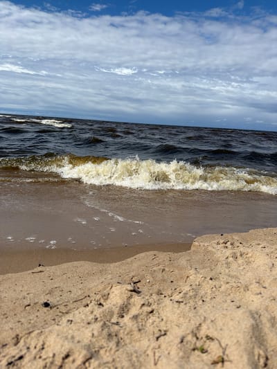 A low-angle, eye-level shot depicts a beach scene in Divezeri, Latvia. The foreground is dominated by a sandy beach, with some small pieces of debris scattered on the sand. A gentle wave with white foam is breaking on the shore, with small bubbles visible on the wet sand. The water is dark and choppy, stretching out to the horizon. The sky above is a mixture of bright blue and scattered white clouds. No people are visible in the image.