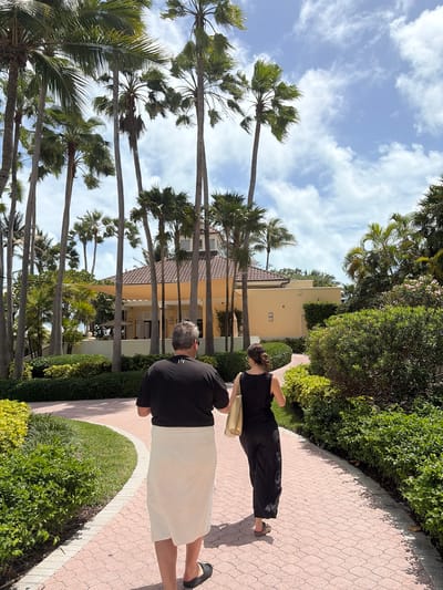 The image depicts an outdoor scene with two people walking along a brick path. A taller man in a black shirt and light-colored shorts walks ahead of a shorter woman wearing a black jumpsuit and carrying a beige bag. The path is bordered by lush green bushes and trees on one side and a row of tall palm trees on the other, leading to a tan-colored building in the background. The sky above is partly cloudy. The location is Key Biscayne, United States of America.