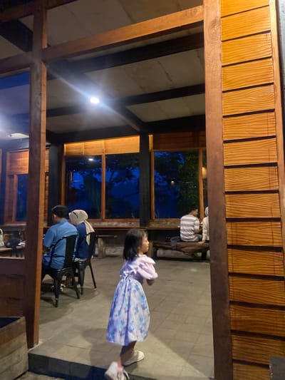 The image depicts a scene at night in what appears to be an open-air structure with a wooden frame and roof. The location may be in Tamiajeng, Indonesia. The roof is constructed with parallel wooden beams. A light fixture is visible on the ceiling.
Several people are present. A girl in a light-colored dress with a floral pattern is standing and looking toward the camera's perspective. Seated at a table are two individuals, one wearing a light blue shirt and another with a white head covering. In the background, another person in a striped shirt is seated.
The scene suggests an informal outdoor dining or gathering area. The lighting is dim. The visible environment includes outdoor vegetation seen through the structure's openings.
