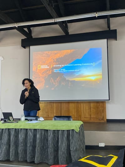 The image shows an indoor setting where a presentation or seminar is taking place. A woman stands in the foreground, facing slightly towards the camera with a subtle smile and her hands clasped. She has dark, curly hair and is dressed in a dark jacket, a blue top, and blue jeans.
Behind her, a large projection screen displays a presentation slide. The slide features a scenic landscape with mountains under a dramatic sky, possibly at sunrise or sunset, with warm, golden light. The "NATIONAL GEOGRAPHIC" logo is prominently displayed in the bottom left corner of the slide. The visible text on the slide reads: "Creating an Inclusive Learning Community," followed by "Shannon P. Bartlett, Chief Diversity, Equity & Inclusion Officer."
To the woman's right, a long table is covered with a dark, pleated fabric and a bright green cloth runner. A laptop and a glass are visible on the table. A dark chair is partially seen behind the table. On the far right, part of a flag, predominantly black with yellow and red shapes, is visible. To the left of the screen, a metal structure resembling scaffolding is present. A wooden cabinet or door is also visible in the background between the screen and the table. The overall scene suggests an educational or corporate event. The provided location context for this event is Riara Ridge, Kenya.