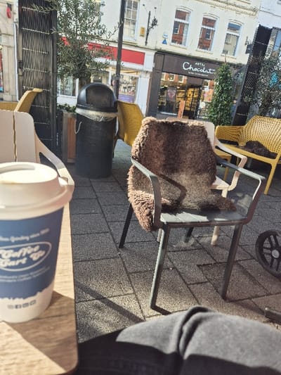 The image captures an outdoor street scene in High Town, Hereford, United Kingdom. In the immediate foreground, a white and blue paper coffee cup labeled "Coffee Cart Co" rests on a light wooden surface, partially obscuring the lower left. The upper thighs and knees of an individual wearing dark trousers are visible in the bottom right corner, suggesting a first-person perspective. The midground features a paved outdoor seating area with grey square tiles, illuminated by sunlight casting distinct shadows. Several unoccupied chairs are present: a dark grey metal chair with a brown, furry cushion in the center-right, and multiple yellow plastic chairs scattered throughout. A black, cylindrical public waste bin with a visible plastic liner is positioned towards the left. Small trees or potted plants with dark foliage are interspersed within the seating area. The background reveals commercial buildings lining a street. Prominently centered is a "Hotel Chocolat" storefront with a dark brown facade and white signage. To its left, a building with a red fascia is partially visible. Above the ground-level shops, white-painted upper stories with multiple windows are observed. No other individuals are present in the visible scene.