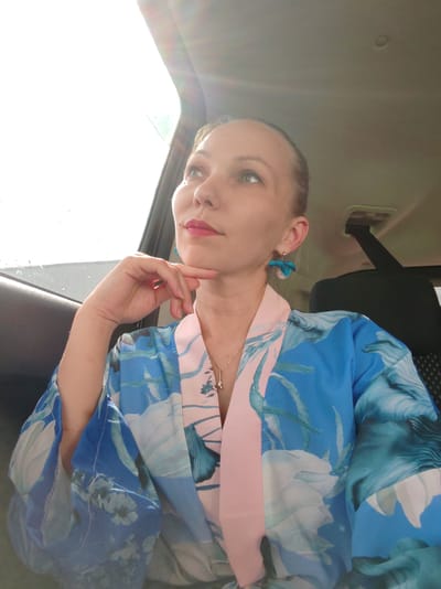A woman with blonde hair pulled back is seated in a car, looking out the window with a thoughtful expression. She is wearing a blue and pink floral robe and turquoise earrings. Her hand is resting on her chin. The sun is shining brightly through the car window, casting lens flares across the image. The interior of the car, including a seatbelt and part of the headrest, is visible. There is no information in the image to suggest this scene is in Syktyvkar, Russia.