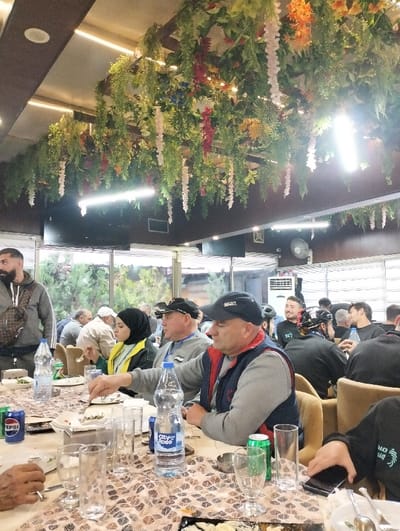 A group of people are seated around a long table, appearing to be dining together in a restaurant setting. The table is covered with dishes, drinks, and glassware. Several individuals are wearing caps and jackets. The ceiling is adorned with a dense arrangement of artificial flowers and greenery, creating a vibrant and festive atmosphere above the diners. Large windows provide natural light and a view of the outdoors.