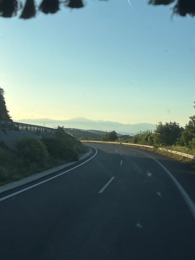 The image depicts a winding asphalt road curving to the right, bordered by a metallic guardrail and lush green vegetation on the left. In the distance, a range of hazy mountains stretches across the horizon under a clear, pale blue sky. The scene appears to be captured from a moving vehicle, as indicated by the blurred dark shapes at the top of the frame, suggestive of a windshield or roofline, and a faint contrail visible in the sky. The lighting suggests either early morning or late afternoon, with a soft, diffused sunlight. The location is identified as Avra, Greece.