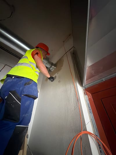 A male construction worker wearing a neon yellow safety vest, orange hard hat, and blue work pants is standing on a wooden ladder. He is holding a silver metal pipe and appears to be working on a wall, possibly installing or repairing ventilation. The environment suggests an indoor construction site, with exposed pipes, wiring, and unfinished walls. The scene is well-lit, with the worker being the primary subject. The overall impression is one of active construction work.