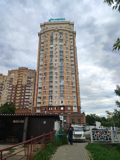 A tall, modern, light brown brick apartment building with a beige facade and numerous windows stands prominently in the center of the frame under a cloudy sky. A green sign with the word "МЕДСИ" is visible at the top of the building. To the left of this building, a slightly shorter, similar apartment building with a brown and beige facade is visible. In the foreground, a paved walkway runs alongside a red metal fence and lush green grass. A man in a dark blue vest and jeans is walking on the walkway, talking on his phone. Several cars are parked along the street, including a white minivan and a black sedan. A covered structure with an open garage door is situated to the left of the walkway, containing wooden pallets. The scene appears to be an urban residential area in Shchyolkovo, Russia, possibly a street or parking area adjacent to the buildings.