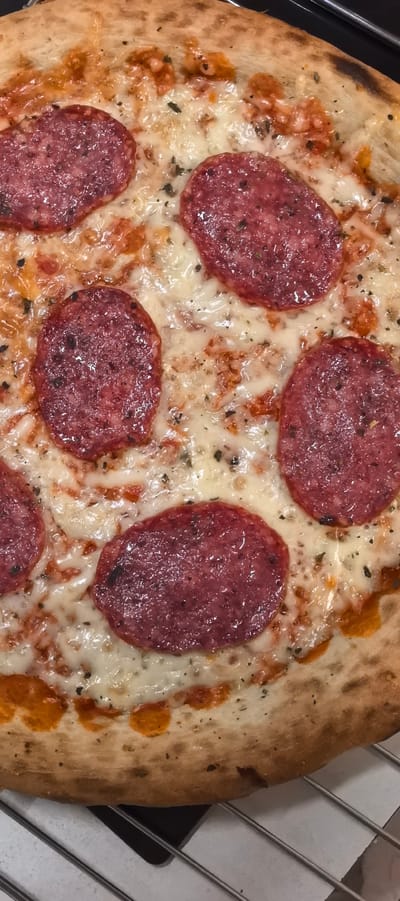 This is a close-up, overhead shot of a pizza. The pizza has a golden-brown, slightly puffed crust. It is topped with melted white cheese, visible red sauce, and several round slices of pepperoni. Small green herbs are sprinkled over the cheese. The pizza is resting on a metal cooling rack, which is partially visible at the bottom of the frame. The background is dark and out of focus. There are no people or identifiable landmarks of Villeneuve-le-Roi, France visible in this image.
