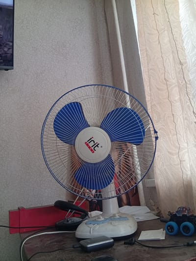 A stationary fan with blue blades and a white center bearing the "irit HOME" logo stands on a wooden surface in front of a patterned wallpaper. To the left of the fan, a red rectangular object is visible. In front of the fan, there are several items, including a black stapler, a white object that appears to be a fan base or decorative element, and a small grey electronic device connected by a black cable. To the right of the fan, a toy monster truck with blue wheels and a colorful body is present, along with white curtains in the background. The scene suggests an indoor setting, likely a room. The presence of the fan and the toy truck indicates a personal space. There are no people or animals visible in the image. The lighting appears to be natural, coming from the window behind the curtains.