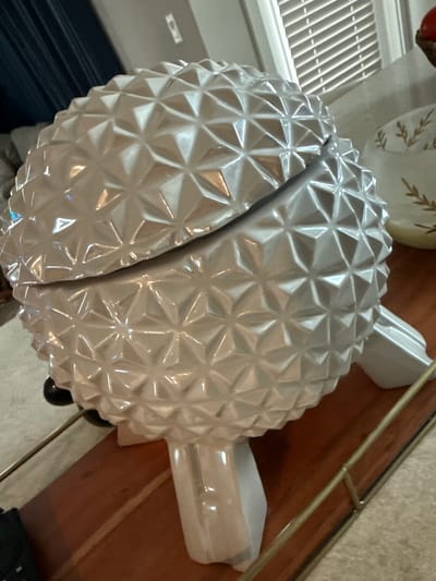 The image shows a silver-colored, decorative object resembling a pineapple sliced in half, sitting on a wooden surface. The pineapple has a textured surface consisting of small, raised triangular shapes. It features stem-like projections on each half. The object is located indoors, near a window with horizontal blinds, and possibly on a table with a metal railing. The location appears to be a residence or commercial building in Orlando, United States.