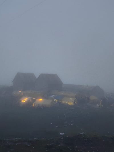 A settlement of tents and some larger structures, possibly buildings, is visible through heavy fog. Several tents are illuminated from within by a warm, yellow light, suggesting occupancy. In the background, two larger, dark, hexagonal structures are silhouetted against the foggy sky. The ground appears uneven and is covered in dark soil and scattered debris. To the far right, a single illuminated figure stands near a small object on the ground. The overall atmosphere is dim and obscured by fog, with a hint of activity from the lit tents. The scene is located in Marhi, India.