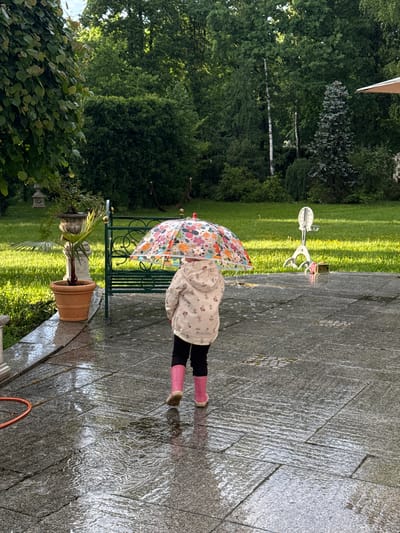 A girl with pink boots and a flowered umbrella stands on a wet, grey stone patio, beside a bench and potted plant. The background shows a green lawn and trees. The scene appears to be in a garden. There is no information to suggest that the location is in the city of Sapronovo, Russia.