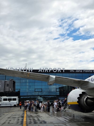 The image shows a group of people boarding an airplane at the John Paul II International Airport Kraków–Balice in Balice, Poland. The airport terminal, with its large glass facade, is visible in the background, with the text "KRAKÓW AIRPORT im. Jana Pawła II" prominently displayed. An airplane wing and engine are in the foreground, obscuring part of the terminal. Passengers, carrying luggage, are forming a line to board the aircraft via a jet bridge. The tarmac is wet, suggesting recent rain, and the sky is cloudy with patches of blue. A service vehicle is parked on the left side of the image, and a person in an orange vest is standing near it. The scene captures a typical airport departure or arrival moment.