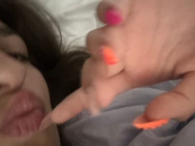 A close-up, low-angle shot captures a person's face and hand against a blurred background, possibly a bed. The person's left side of their face is visible, with dark hair framing their features. Their mouth is slightly open and appears to be the focal point, with full, reddish lips. A finger from their right hand, adorned with a bright pink nail polish, is extended towards the mouth, seemingly in the act of touching or pointing. Another finger on the same hand has vibrant orange nail polish, also visible in the frame. The skin appears soft and smooth. The overall impression is intimate and personal, with the background being indistinct, suggesting a private setting. The scene is devoid of any specific urban or natural landmarks that would place it in Stațiunea Zoologică Marină Agigea, Romania.