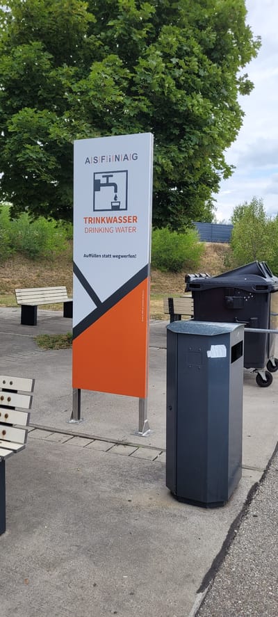 The image depicts an outdoor scene in Pöchlarn, Austria, near the Danube River. In the foreground, a dark gray, hexagonal trash receptacle stands on a concrete surface. To the left, a light-colored bench with a slatted seat and black legs is visible.
Dominating the center of the image is a tall, white sign with an orange and black triangular graphic at the bottom. The top of the sign features text and a symbol: "A|S|F|i|i|N|A|G" is written across the top, followed by a black outline of a faucet with a droplet of water falling into a cup. Below this, the text "TRINKWASSER" (German for drinking water) is in orange, and "DRINKING WATER" is in smaller black text. The phrase "Auffüllen statt wegwerfen!" (Refill instead of throw away!) is also present. A small inscription in the lower right corner of the orange section reads "Ich bin zu 100% aus recyceltem Aluminium!" (I am 100% recycled aluminum!).
In the background, another bench is partially visible, and a large, black wheeled trash bin stands to the right of the sign. Trees with lush green foliage form a backdrop, and a gray fence or barrier can be seen in the distance. The ground is paved with concrete slabs and gravel. The sky is overcast with scattered clouds.