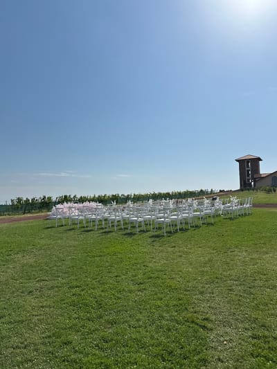 An outdoor scene in Sasunik, Armenia, is depicted on a clear, sunny day. The foreground consists of an expanse of well-maintained green grass. In the midground, multiple rows of white cross-back chairs with white seat cushions are arranged on the grass, facing towards the left side of the frame. To the immediate left of the chair setup, a structure adorned with light pink and white floral decorations is visible, positioned as a potential arch or backdrop. The background on the left features rows of grapevines, indicative of a vineyard, extending horizontally across the landscape. A dirt path runs parallel to the vineyard rows. On the right side of the background, a large stone building with a prominent square tower topped by a red-tiled roof stands on a slight elevation. The tower exhibits multiple arched openings. The sky overhead is a uniform bright blue, devoid of clouds. No individuals are present in the scene.