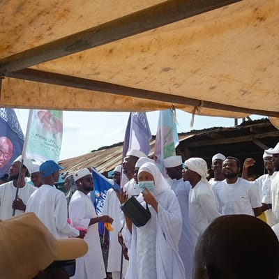 Captured under an open-air, tarpaulin-covered shelter in Gudi, Nigeria, the image features a crowd of people primarily dressed in white. Some are wearing caps, head wraps, or turbans, while one individual is wearing a blue face mask and carrying a black handbag. Several are holding flags with political messages. The background includes a partially visible corrugated iron roof structure. The composition is a medium shot, capturing the subjects from various angles within the crowd.