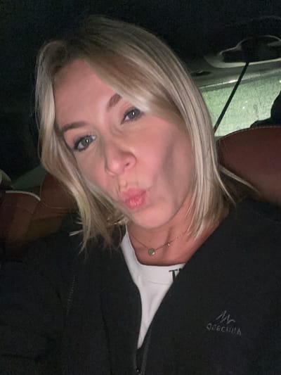A blonde woman with blue eyes poses for a selfie. She is wearing a black jacket over a white t-shirt and a delicate gold necklace. Her lips are puckered. The background is dark, with a hint of a car interior visible in the upper left corner. The picture appears to be taken in low lighting.