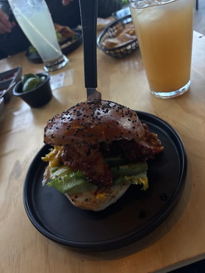 The image shows a table with a black plate containing a burger. The burger appears to be cut open with a knife, revealing lettuce and possibly other fillings. A glass of orange juice is present to the left of the plate. Other items, including what seems to be a small container of limes and a utensil, are also partially visible on the table. The location is San Pedro Cholula, Mexico.