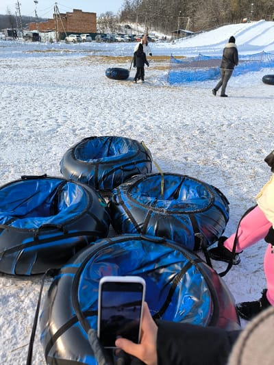 The image depicts a winter scene at a snow tubing facility in Prybuzke, Ukraine. In the immediate foreground, four black and blue snow tubes are visible, arranged on packed snow. A person's hands, gloved in black, hold a white smartphone positioned over one of the tubes, capturing a horizontal image of a snowy landscape. To the right of the foreground, the lower torso and legs of another person are visible, wearing bright pink snow pants and black boots, standing near the snow tubes.
In the mid-ground, a child in dark winter clothing pulls a single black and blue snow tube across the snow-covered ground. An adult in a white jacket stands near the child. Further to the right, another adult, dressed in a dark jacket and black hat, walks across the snowy terrain, moving past a section of blue safety netting or fencing. Another snow tube is visible near this walking adult.
The background features a snow-covered hill or slope on the right. To the left, several multi-story brick-colored buildings are visible, beyond which a parking area contains numerous parked cars and SUVs. Utility poles with overhead lines are present. The overall environment is bright, suggesting daytime conditions with a clear sky, and the ground is consistently covered in snow.