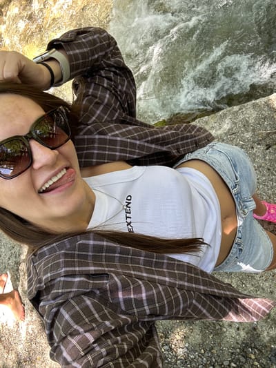 A young woman with long, brown hair is lying on her back next to a body of water, possibly a stream or river. She is wearing a white tank top with the word "EXTEND" printed on it, denim shorts, a brown and purple plaid button-up shirt worn open, and pink shoes. She is also wearing sunglasses and a watch on her left wrist. Her tongue is sticking out and her mouth is open. The water appears to be moving and shallow, with rocks and pebbles visible in and around it. The ground beneath her is rocky and sandy. The overall impression is a casual, outdoor scene. The location is not specified in the image or description.