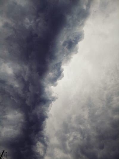 This is a photograph of a dramatic sky filled with dark, ominous storm clouds. The clouds dominate the frame, with a stark division between the deep gray, almost black, clouds on the left and lighter, white-gray clouds on the right. The texture of the clouds suggests turbulence and the imminent threat of rain or a storm. There are no visible people or structures in the immediate foreground or background, but the extreme left edge of the image shows a small portion of a dark, angular object that could be part of a man-made structure, possibly a power line or tower, silhouetted against the sky. The overall impression is one of natural power and impending weather change. The image is a close-up view of the sky, providing no context for its specific location within Tash-Dobo, Kyrgyzstan.
