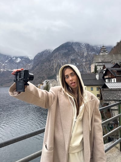 A female individual is depicted outdoors, posing for a self-portrait or video recording with a digital camera held in her extended left hand. She has long, dark brown hair and is wearing a light beige oversized coat over a cream-colored zippered hoodie and matching cream sweatpants. Her eyes are wide, and her tongue is extended from her mouth. The camera, black in color with an articulated screen facing the subject, is held at shoulder height.

The scene is set in Hallstatt, Austria, alongside Hallstätter See (Lake Hallstatt). Two parallel grey metal railings are visible in the immediate foreground, separating the subject from the dark, calm lake water to her left. In the mid-ground, across the lake, traditional Alpine houses with distinctive roofs are visible, nestled against the base of steep, rocky mountains. The prominent spire of St. Michael's Church rises above the houses on the right side of the frame. Further up the hillside, another structure with a domed tower is visible. The mountains, partially covered with snow and sparse vegetation, recede into the background under an overcast, light grey sky with clouds obscuring some peaks.