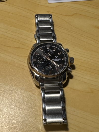 A silver-toned Daniel Klein Exclusive wristwatch is placed on a light brown wooden surface. The watch has a black dial with silver hour markers and hands. There are three sub-dials on the main dial, indicating days of the week, 24-hour time, and seconds. The watchband is made of interconnected silver links. The background appears to be a wooden table or desk. No people or any other objects are visible in the frame. The scene does not provide any information about the city of Sharjah, United Arab Emirates.