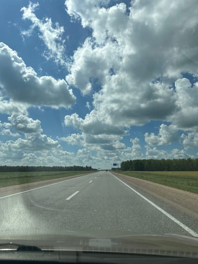 The image shows a view from a car driving on a straight asphalt road in Қызылжар ауылдық округі, Kazakhstan. The road has a white dashed line marking the lanes and a solid white line on the right edge. To the left of the road, there is a grassy field bordering a dense line of dark green trees. The sky is filled with large, fluffy white and gray clouds against a bright blue sky, suggesting a daytime setting with partly cloudy weather. The perspective is from inside the car, with a portion of the car's interior visible on the right side of the frame, indicating the photograph was taken while in transit. There are no people clearly visible, but a few small distant objects on the road may be other vehicles. The scene conveys a sense of travel through a rural landscape under a dynamic sky.