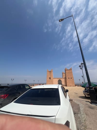 The image depicts an exterior shot of the Kasbah of Beni Mellal in Beni Mellal, Morocco, under a blue sky with scattered clouds. The Kasbah is a brick-colored building with castle-like features. Two cars, one white and one dark gray, are parked on the adjacent road. Street lamps line the road. People are visible in the distance, near the Kasbah. Part of a green umbrella is visible at the top of the frame. A tall light pole extends from the foreground toward the sky.