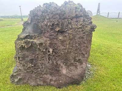 A large, rough, dark gray volcanic rock with many holes and crevices sits in a grassy park in Kuril'sk, Russia. The rock is the main subject, taking up the majority of the foreground. The background is a hazy, overcast sky with a green fence and a lamppost visible to the left. To the right, a hill with a metal sculpture and a decorative fence can be seen. The ground is covered in bright green grass, with small gray pebbles around the base of the rock. The weather appears to be foggy or misty, as indicated by the lack of sharp details in the background. There are no people in the image.