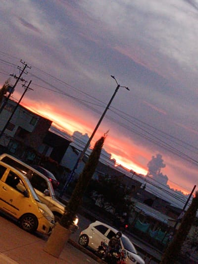A vibrant sunset fills the sky with hues of orange and red behind buildings and utility poles. Streetlights stand tall amidst the urban landscape. Several cars, including a yellow taxi, are parked along the roadside. A motorcycle rider wearing a mask is present near a white car. Tall, slender trees in planters add a touch of greenery to the scene. The overall ambiance is one of urban life bathed in the warm glow of twilight.