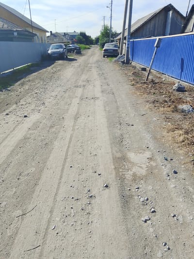 The image shows a dirt road in a residential area of Iskitim, Russia. The road is unpaved and appears to be in poor condition with many small stones and tire tracks visible. On the left side of the road, there is a light-colored fence and a building with a dark roof. Further down the road, several cars are parked. On the right side of the road, there is a bright blue metal fence and a wooden building with a dark roof. The sky is clear and blue, suggesting a sunny day. The scene depicts a rural or semi-rural environment typical of a Russian provincial town.