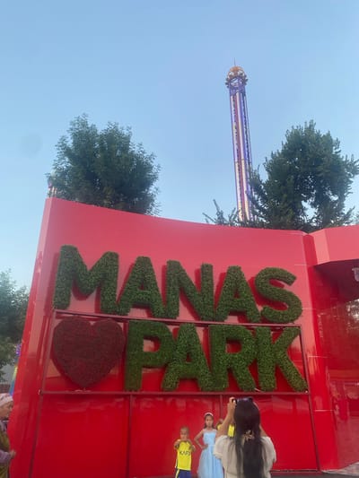 A tall, slender amusement park ride, illuminated with colorful lights, stands against a clear blue sky. To the right, a large, bright red building features the word "MANAS PARK" spelled out in large, green, topiary letters. Below the letters, a heart-shaped topiary adds a decorative element. Several people are visible near the building. A woman with long dark hair is taking a photo with her phone. Two children, one in a blue dress and another in a yellow shirt with the word "KAFD" visible, are also present. The scene appears to be set during the daytime, with bright sunlight. The presence of the amusement park ride and the building signage suggests a theme park or entertainment venue.